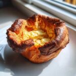 A single, golden brown, and deeply puffed yorkshire pudding sitting on a white plate near a window.