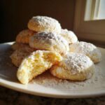 A stack of soft, powdered sugar-dusted cool whip cookies on a white plate, one broken open to show the texture.