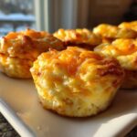 A close-up of several golden, cheesy protein egg bites served on a white rectangular plate near a window.