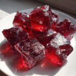 Close-up of several deep red, jagged pieces of cinnamon rock candy piled on a white plate, catching the sunlight.