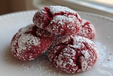 Four rich red velvet cake mix cookies dusted heavily with white powdered sugar, stacked on a white plate.