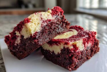 Two rich, moist slices of red velvet cheesecake brownies with a prominent cream cheese swirl, sitting on a white plate.