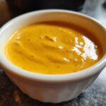 Close-up of smooth, vibrant orange beer cheese dip served in a small white ramekin.