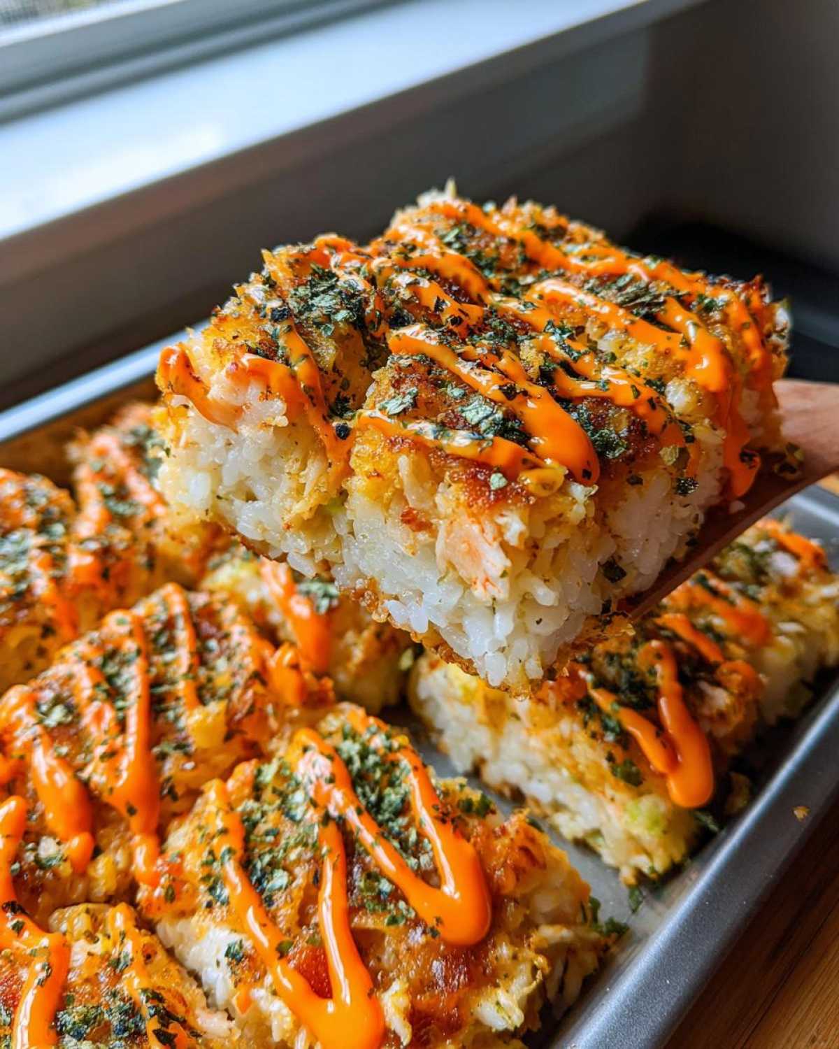A square portion of baked rice topped with spicy mayo and furikake being lifted from a pan of sushi bake.