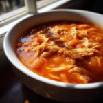 Close-up of a white bowl filled with vibrant orange slow cooker buffalo chicken chili, featuring shredded chicken and beans.
