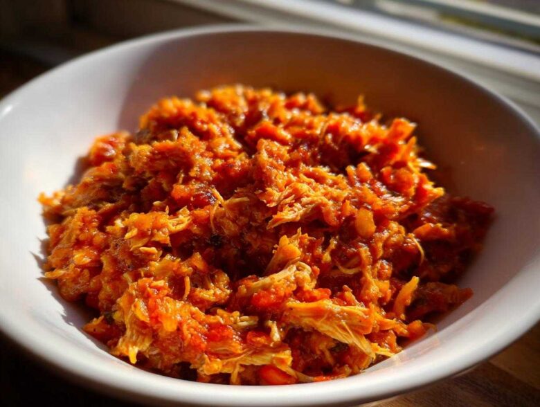 Close-up of shredded chicken in a vibrant orange sauce, representing the slow cooker buffalo chicken chili, served in a white bowl.