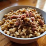 A close-up of a white bowl filled with creamy black eyed peas topped with shredded smoked ham.