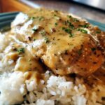 A close-up of smothered chicken and rice, featuring a creamy, seasoned gravy over a chicken breast resting on white rice.