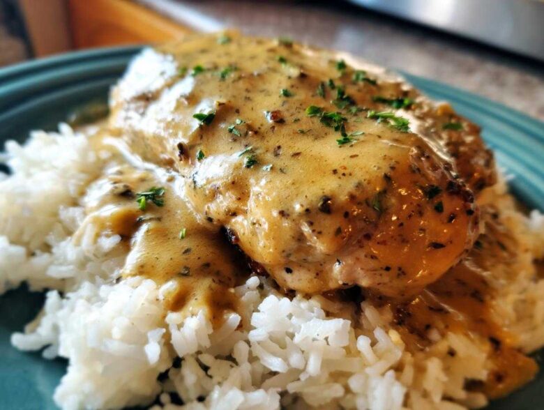 A close-up of smothered chicken and rice, featuring a creamy, seasoned gravy over a chicken breast resting on white rice.