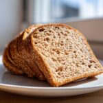 Close-up of several soft slices of honey whole wheat bread stacked slightly on a white plate, catching sunlight.