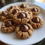 A close-up of several soft peanut butter blossoms cookies, rolled in sugar and topped with a milk chocolate kiss, arranged on a white plate.