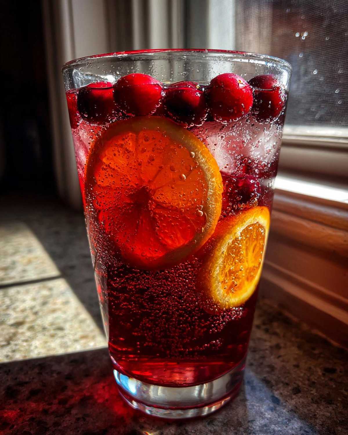 Close-up of a tall glass filled with vibrant red, bubbly thanksgiving punch garnished with fresh cranberries and orange slices.