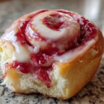 A close-up of a single, soft Strawberry Cheesecake Sweet Rolls, drizzled with white icing and filled with bright red strawberry mixture.