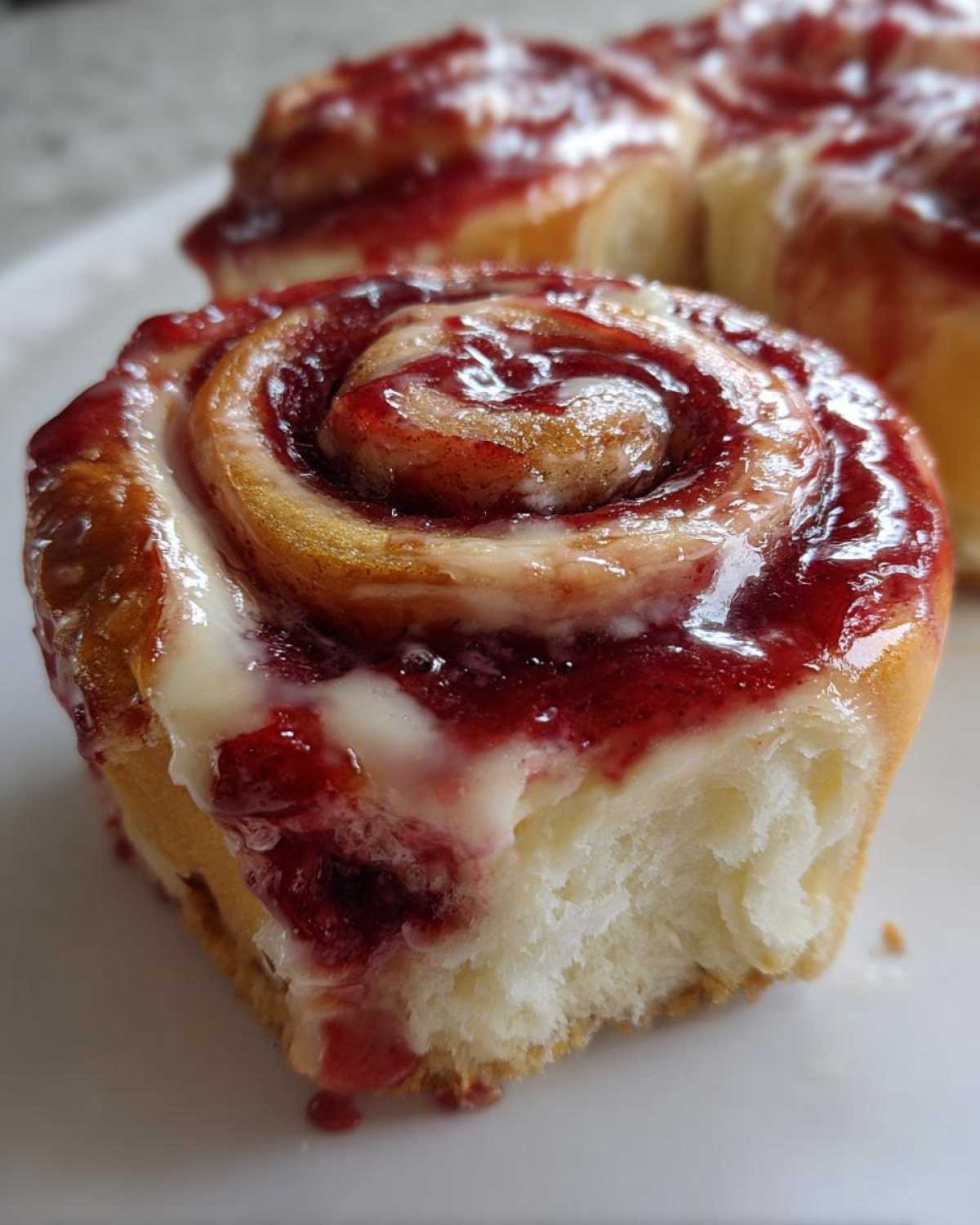 A close-up of a soft, fluffy Strawberry Cheesecake Sweet Roll topped with bright red strawberry filling and white cream cheese glaze.