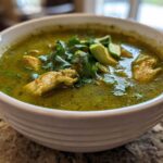 Close-up of a white bowl filled with bright green chicken poblano soup, topped with avocado and cilantro.