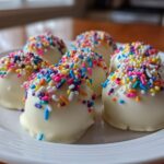 Close-up of several white chocolate sugar cookie truffles topped with colorful rainbow sprinkles on a white plate.