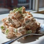 A mound of creamy salmon salad with visible chunks of celery and dill garnish on a white plate.