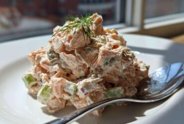 A mound of creamy salmon salad with visible chunks of celery and dill garnish on a white plate.