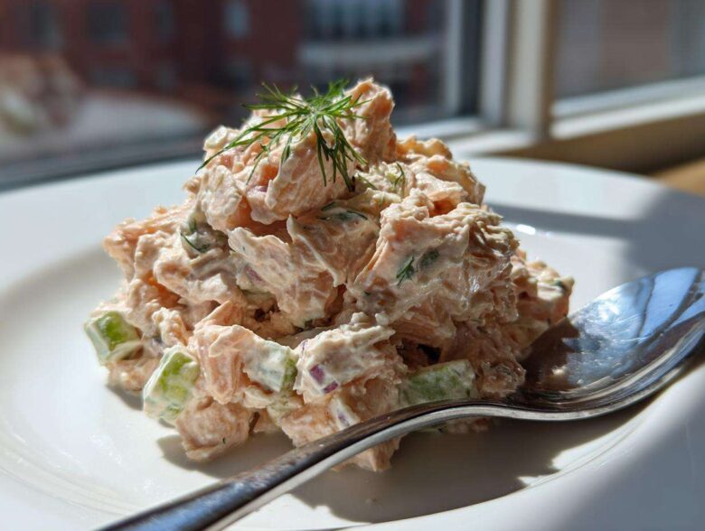 A mound of creamy salmon salad with visible chunks of celery and dill garnish on a white plate.