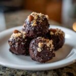 A close-up of several rich, dark chocolate almond joy truffles topped with chopped nuts and whole almonds.