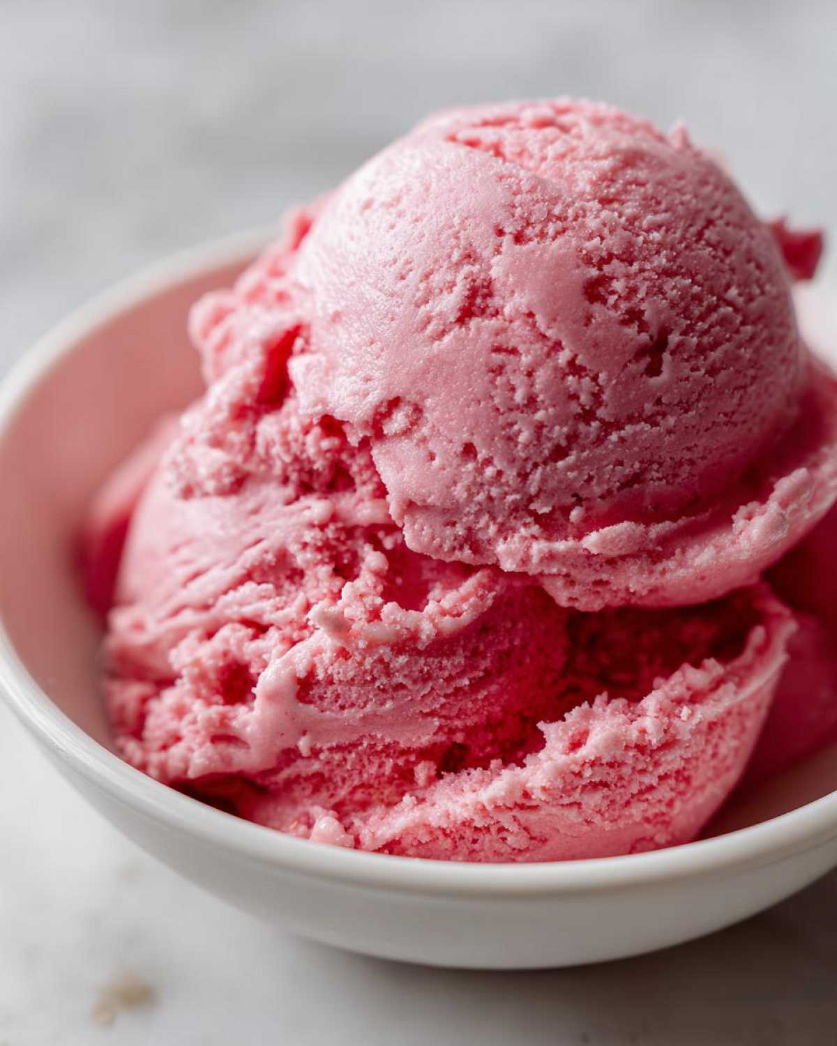 Close-up of two scoops of vibrant pink strawberry ice cream served in a small white bowl.