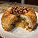 Close-up of a baked brie wrapped in flaky pastry, cut open to show the melted cheese and pecan topping.