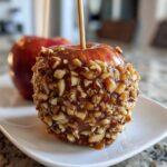 Close-up of a glossy caramel apple coated in chopped nuts, served on a white dish, featuring another apple in the background.
