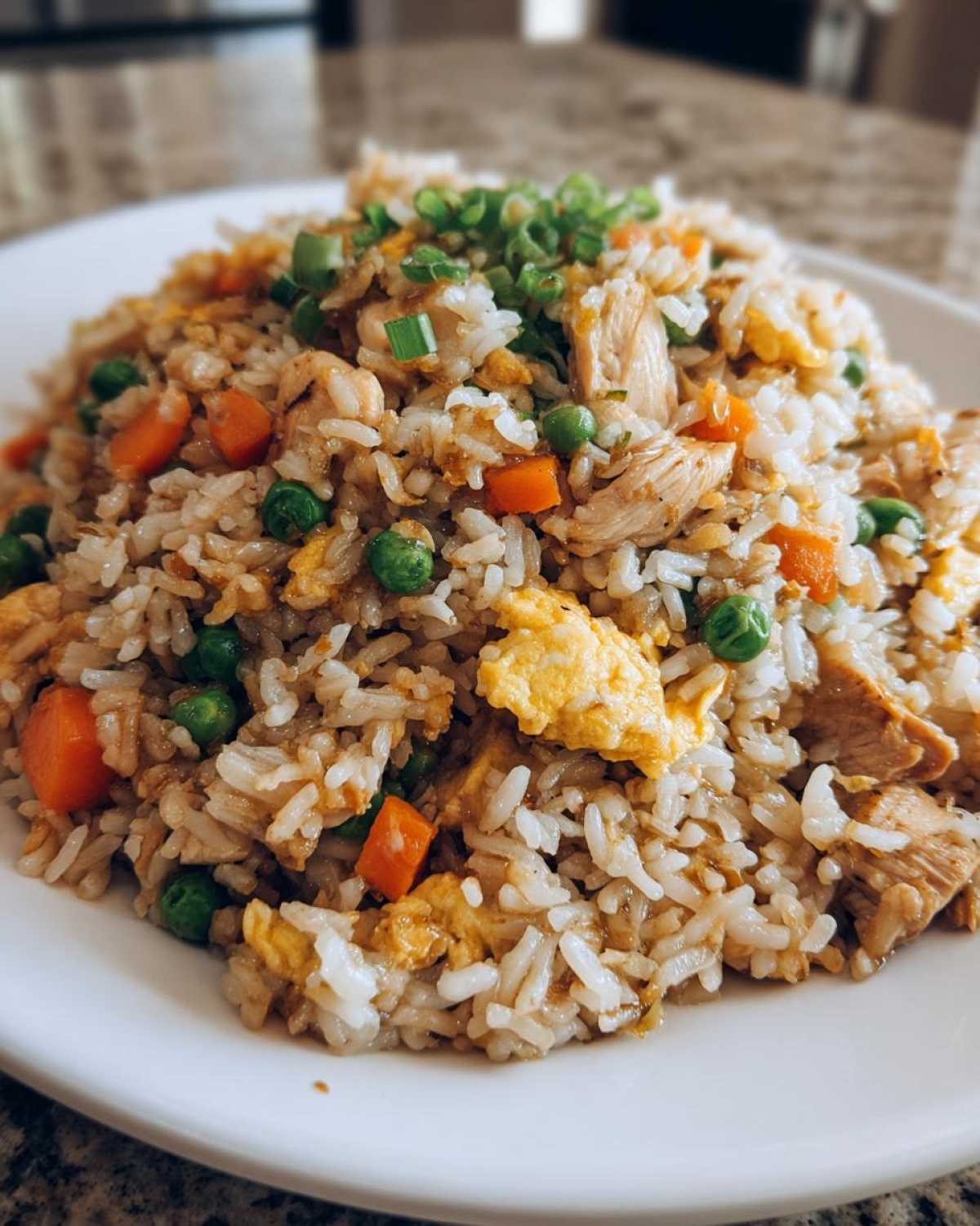 A close-up serving of amazing chicken fried rice mixed with scrambled egg, peas, carrots, and topped with green onions.