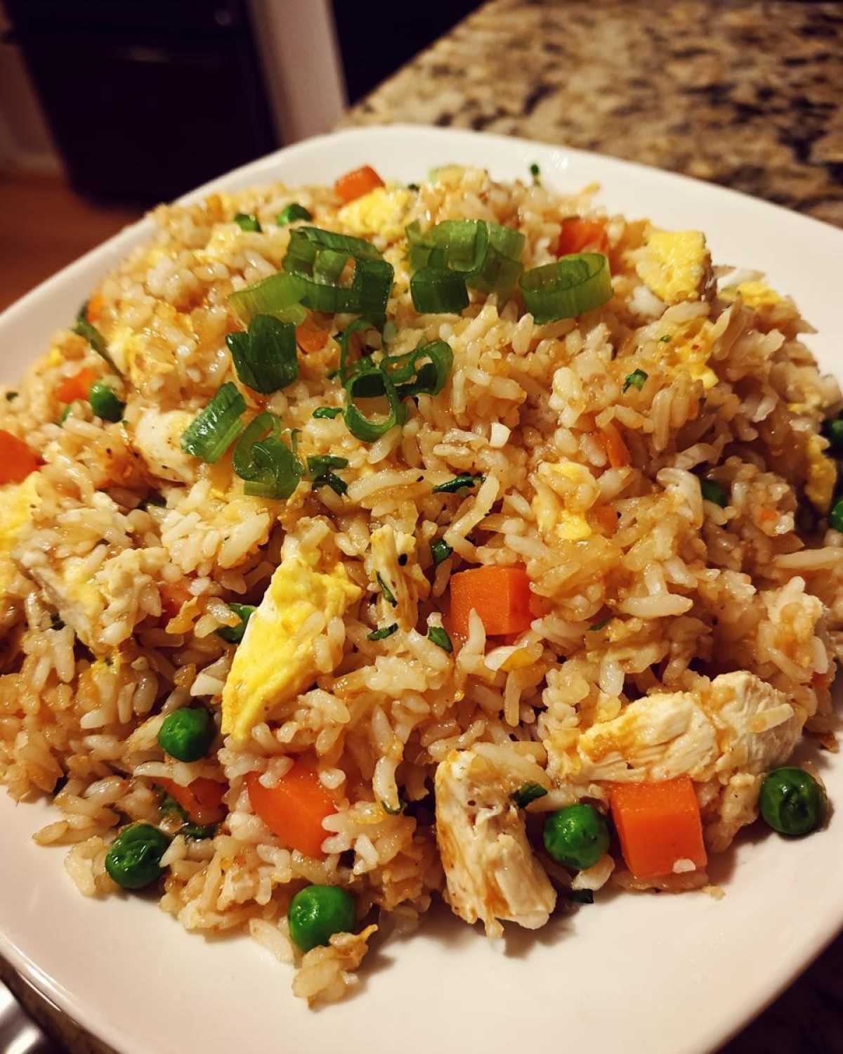 Close-up of a heaping plate of homemade chicken fried rice with visible pieces of chicken, carrots, peas, and egg, topped with green onions.