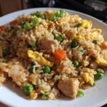 A close-up of a generous serving of homemade chicken fried rice, featuring rice, chunks of chicken, scrambled egg, peas, and carrots.