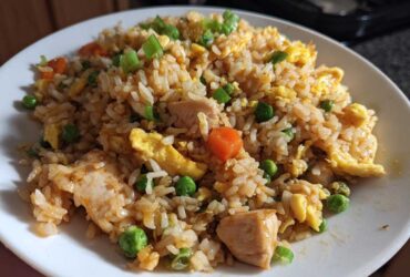 A close-up of a generous serving of homemade chicken fried rice, featuring rice, chunks of chicken, scrambled egg, peas, and carrots.