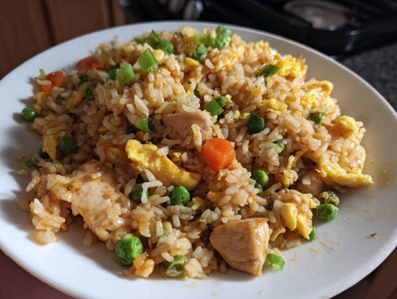 A close-up of a generous serving of homemade chicken fried rice, featuring rice, chunks of chicken, scrambled egg, peas, and carrots.