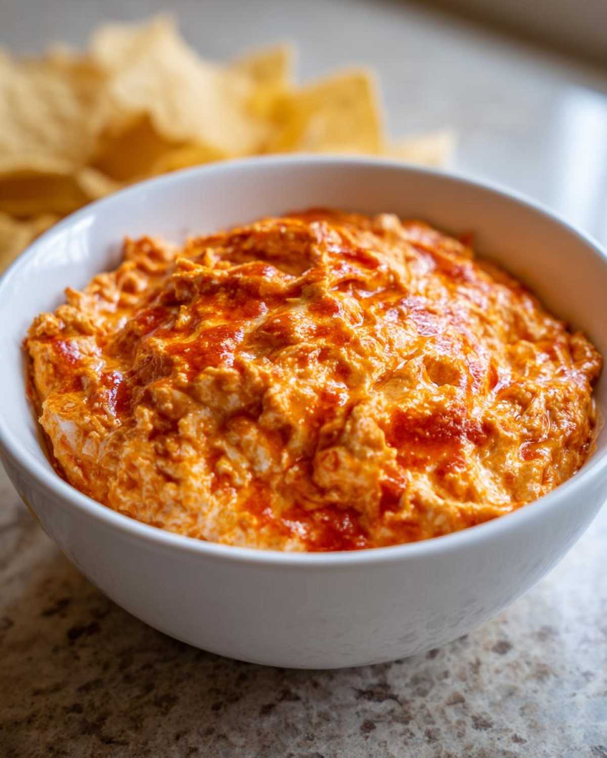 A close-up of creamy, orange chili cheese dip swirled with red sauce in a white bowl, with tortilla chips blurred in the background.