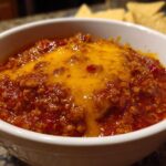Close-up of a white bowl filled with rich, red chili cheese dip topped with melted orange cheddar cheese, with tortilla chips blurred in the background.