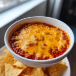 A close-up of a white bowl filled with bubbling chili cheese dip topped with melted orange cheddar cheese, served with tortilla chips.