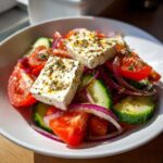 Close-up of a vibrant greek salad featuring thick blocks of feta cheese, ripe tomatoes, cucumbers, and red onion slices.