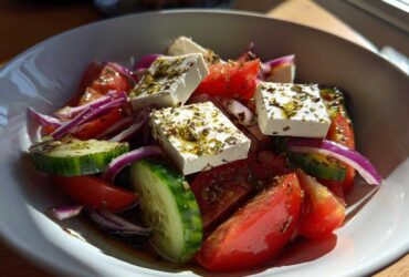 Close-up of a vibrant greek salad featuring large chunks of feta cheese, tomatoes, cucumbers, and red onion drizzled with oil and herbs.
