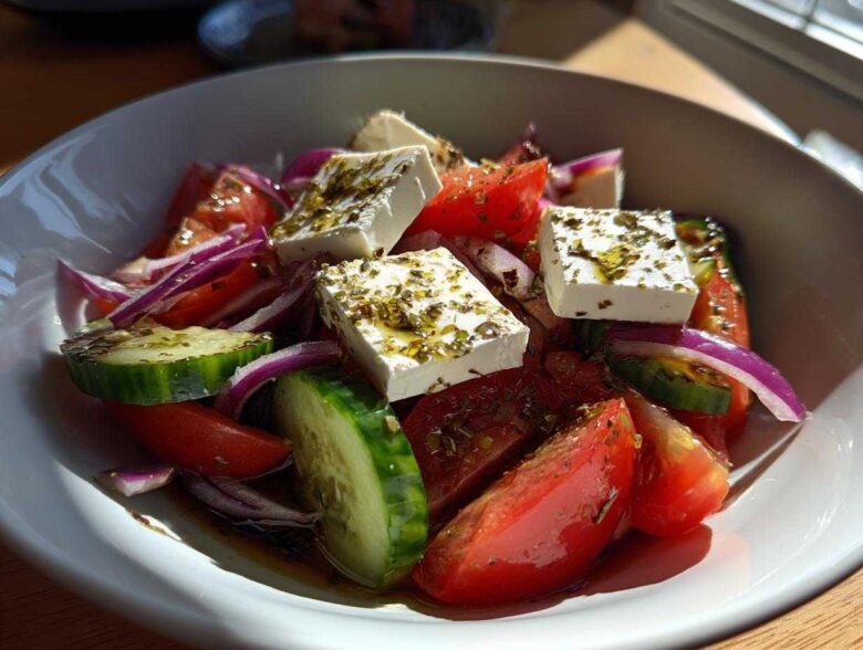 Close-up of a vibrant greek salad featuring large chunks of feta cheese, tomatoes, cucumbers, and red onion drizzled with oil and herbs.