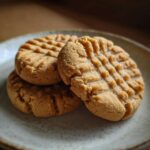 Three freshly baked peanut butter cookies with classic crosshatch fork marks stacked on a speckled ceramic plate.