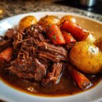 Close-up of shredded pot roast served in rich gravy with roasted carrots and whole potatoes.