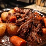 Close-up of shredded, tender pot roast served with whole carrots and potatoes in a rich brown gravy.
