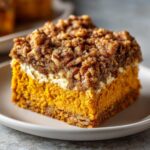 Close-up of a layered slice of pumpkin crunch with a bright orange pumpkin layer, cream cheese layer, and pecan crumble topping.