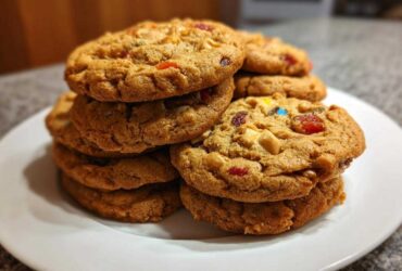 A close-up stack of chewy fruitcake cookies loaded with colorful dried fruit and nuts, featured prominently on a white plate.