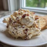 A scoop of creamy Neiman Marcus dip topped with bacon pieces, served on a white plate next to round crackers.