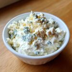 Close-up of a white bowl filled with creamy base topped generously with large crumbles of blue cheese dip.