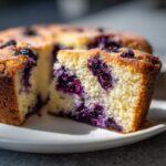 Close-up of a moist slice of blueberry breakfast cake showing bright purple berries embedded in the yellow crumb.