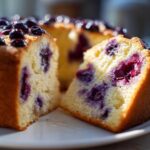 A close-up of a moist slice cut from a blueberry breakfast cake, showing the yellow crumb and dark purple berries.