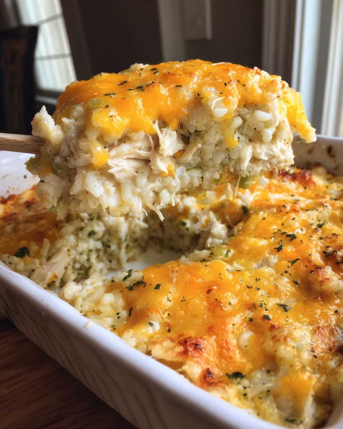 A wooden spoon lifts a generous scoop of creamy chicken rice casserole topped with melted cheddar cheese.