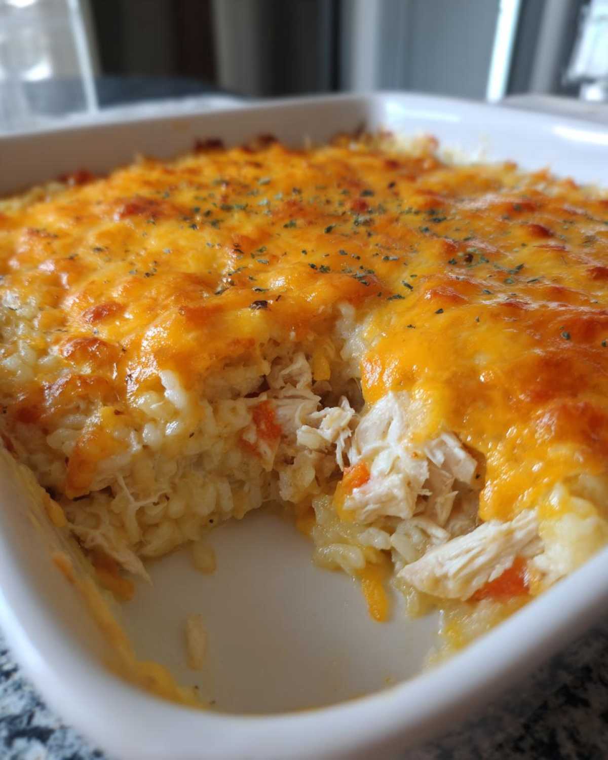A scoop has been taken from a baked rice casserole topped with melted cheddar cheese and herbs.