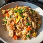 A close-up of a serving of delicious chicken fried rice, featuring chunks of chicken, scrambled egg, peas, and carrots, garnished with green onions.